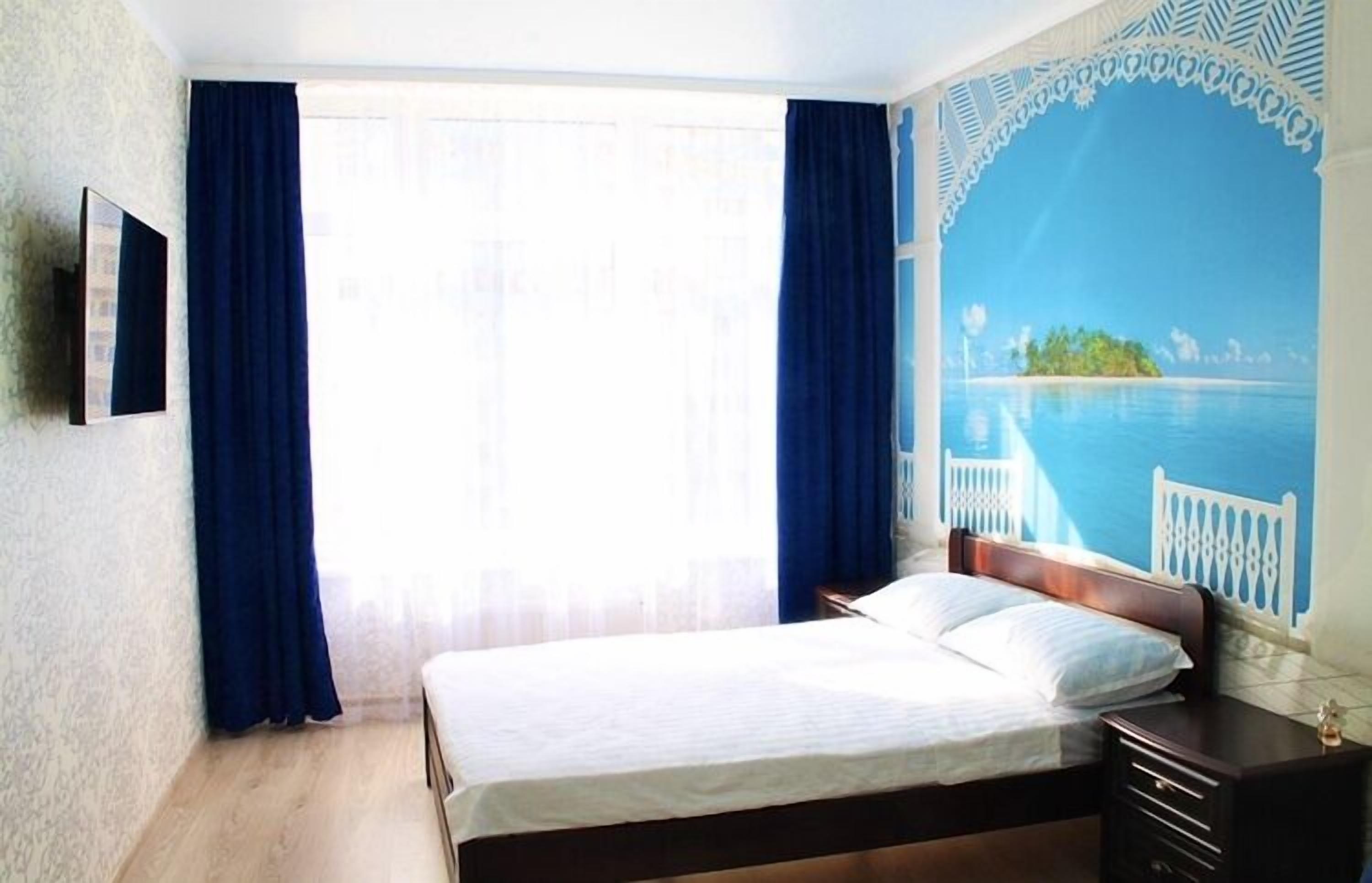 A bedroom with a bed, a nightstand, a TV mounted on the wall, and a mural of a tropical scene.
