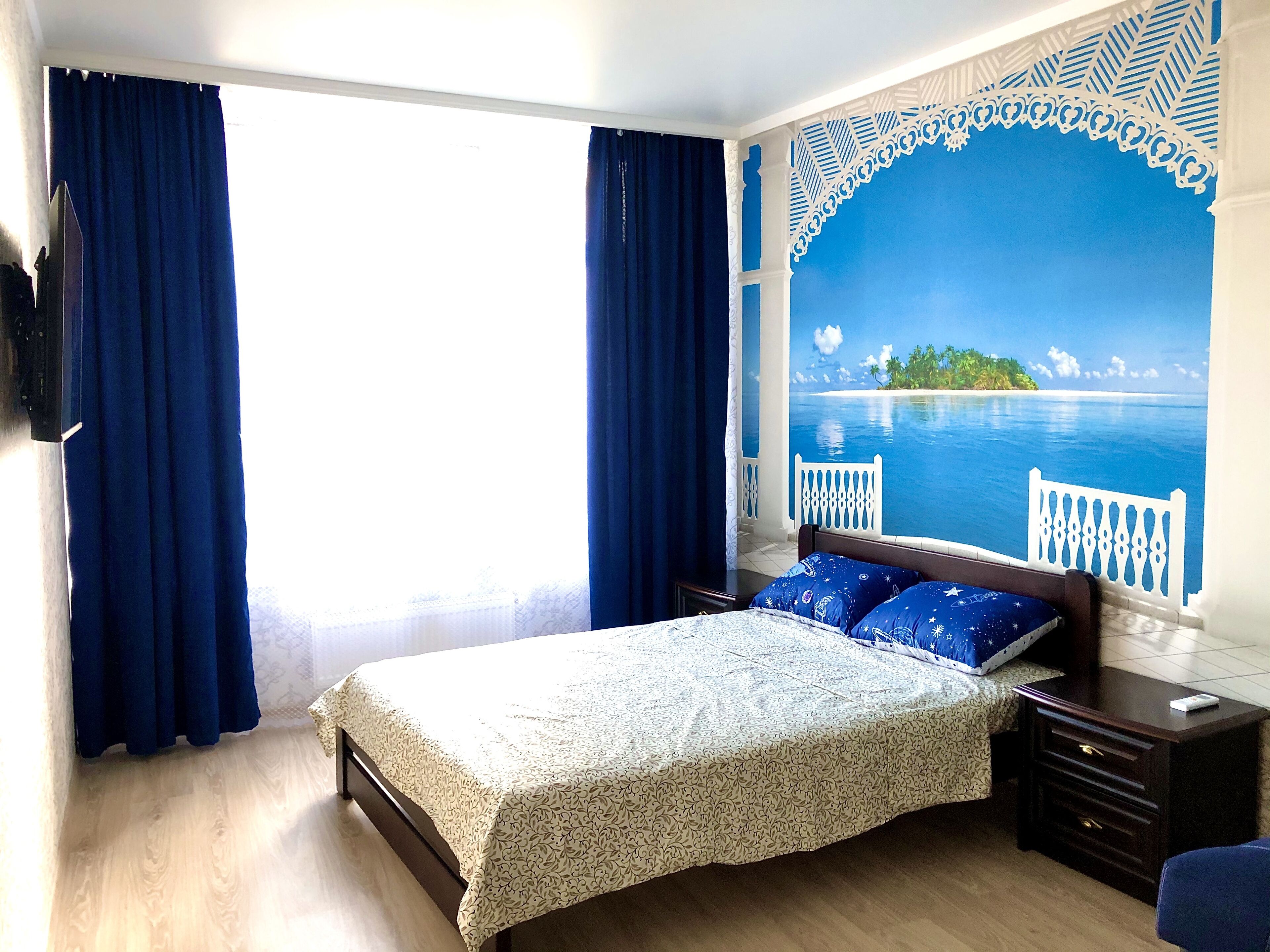 Ein Schlafzimmer mit Bett, Nachttisch, Kommode und einem großen Fenster mit Vorhängen.