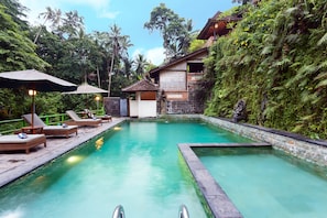 A swimming pool with lounge chairs and umbrellas, surrounded by lush greenery and a stone wall.