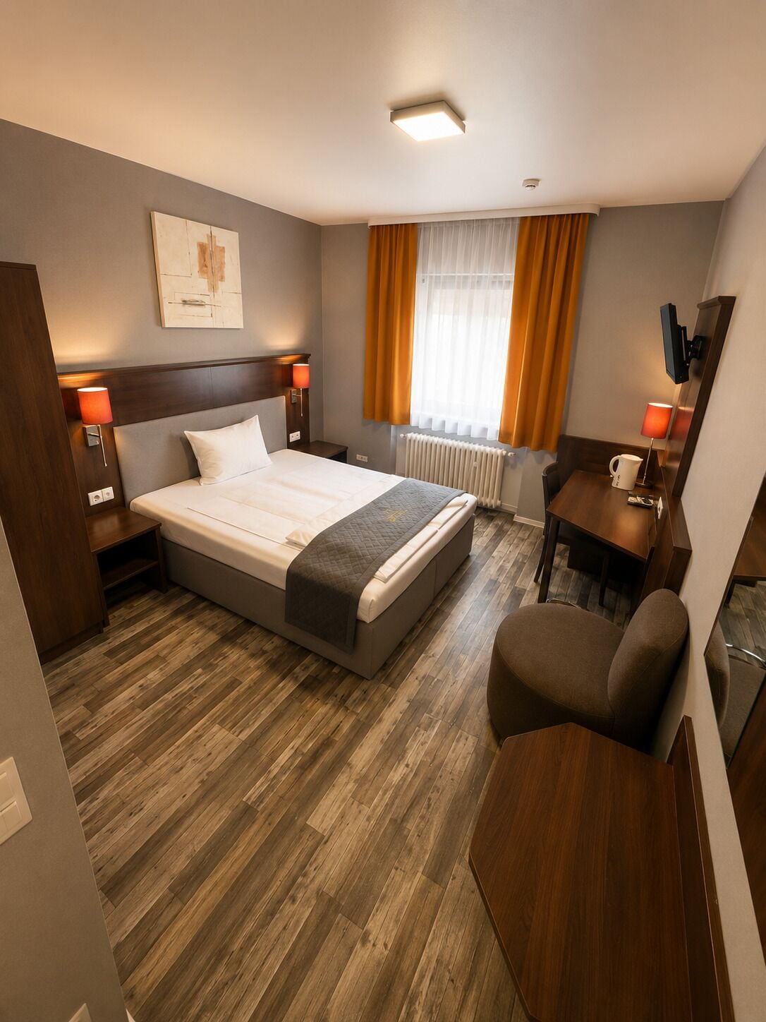 Habitación de hotel con cama, mesitas de noche, escritorio, silla, radiador y ventana con cortinas.
