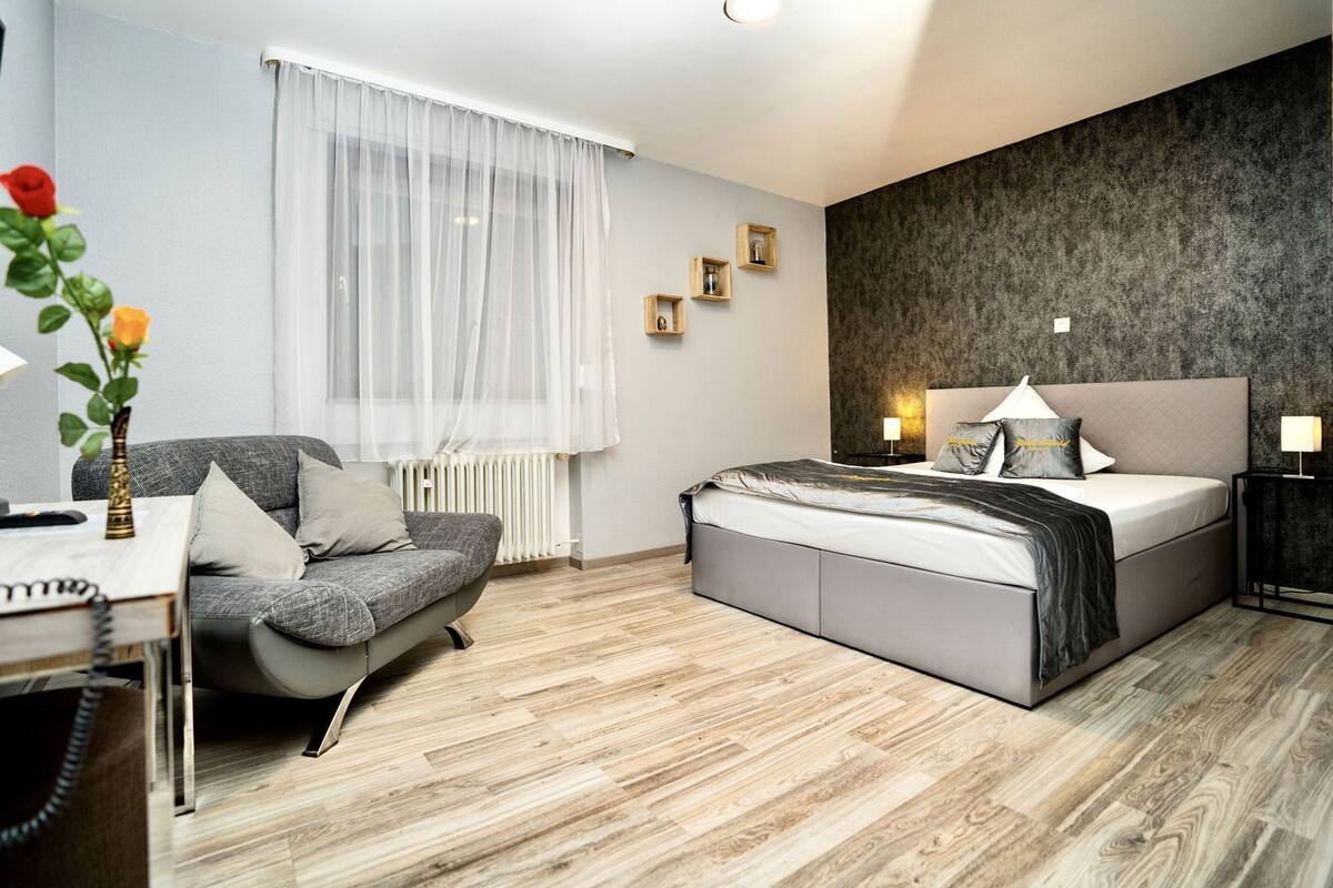 A modern bedroom with a grey sofa, a bed with a grey headboard, a bedside table with a lamp, and a window with sheer curtains.