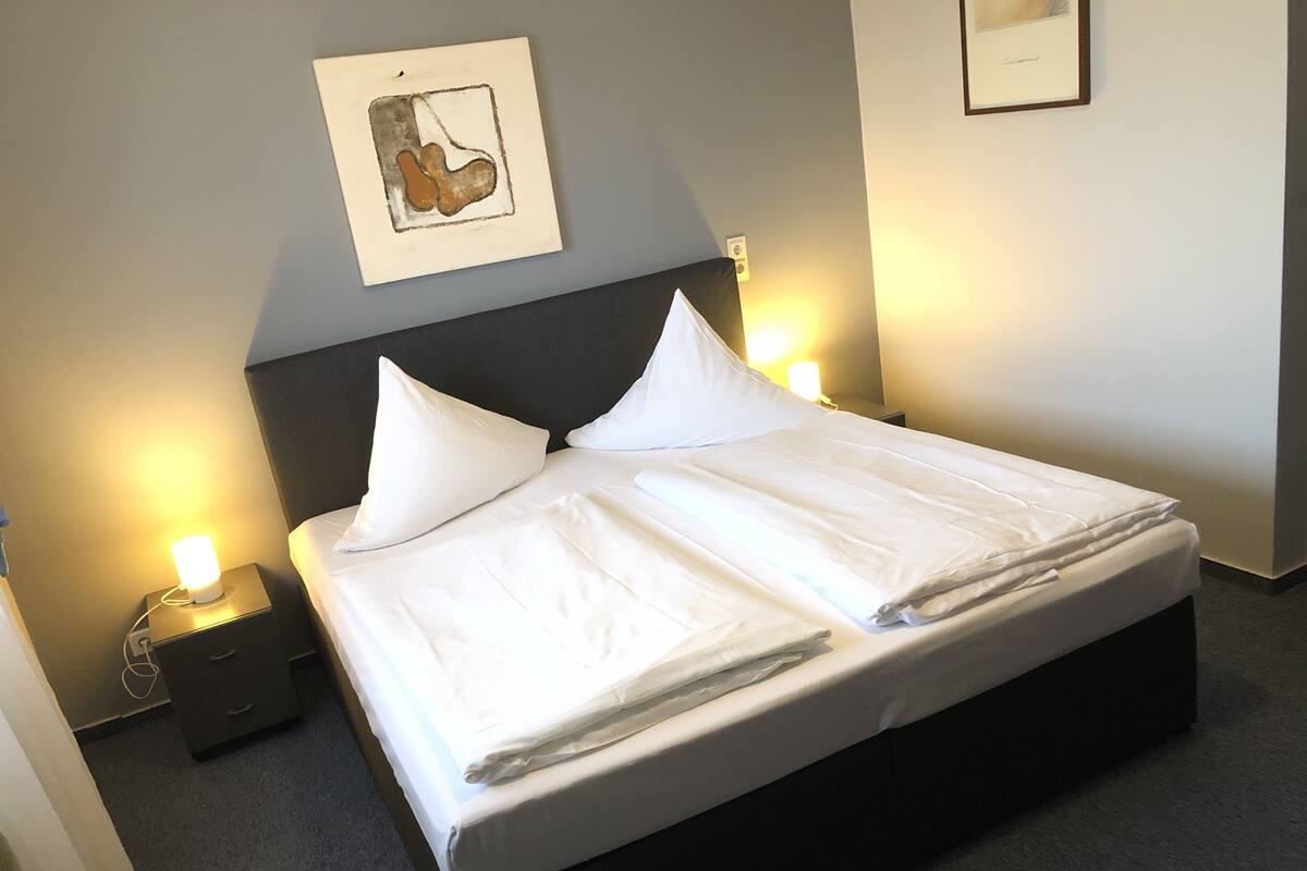 A neatly made bed with white linens, two bedside lamps, a nightstand, and a framed picture on the wall.