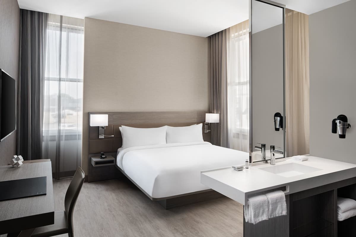 Habitación de hotel moderna con una cama grande, un escritorio con silla, un baño con lavamanos y espejo, y un televisor en la pared.
