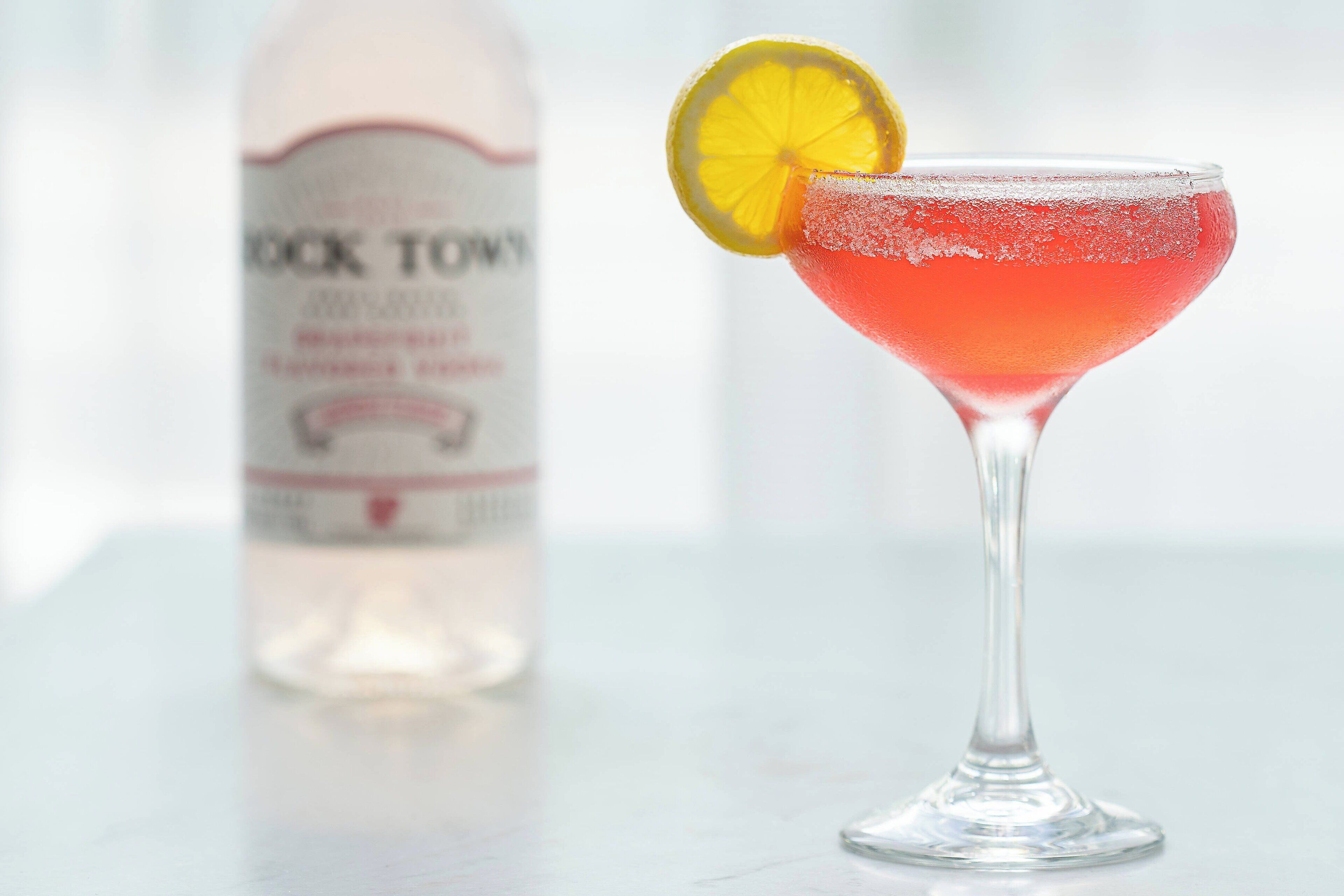 A cocktail in a martini glass with a lemon slice garnish, a bottle labeled "COCK TOWN" in the background.
