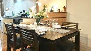A dining area with a wooden table set for four, surrounded by chairs. A vase with flowers is on the table, and there are wine glasses and plates. The kitchen is visible in the background with a microwave and other appliances. - Osaka Premium House (Osaka)