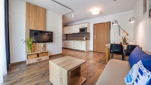 A modern living room with a wooden coffee table, a blue sofa with a nautical design, a flat-screen TV mounted on a wooden wall, and a kitchen area with a microwave and cabinets.