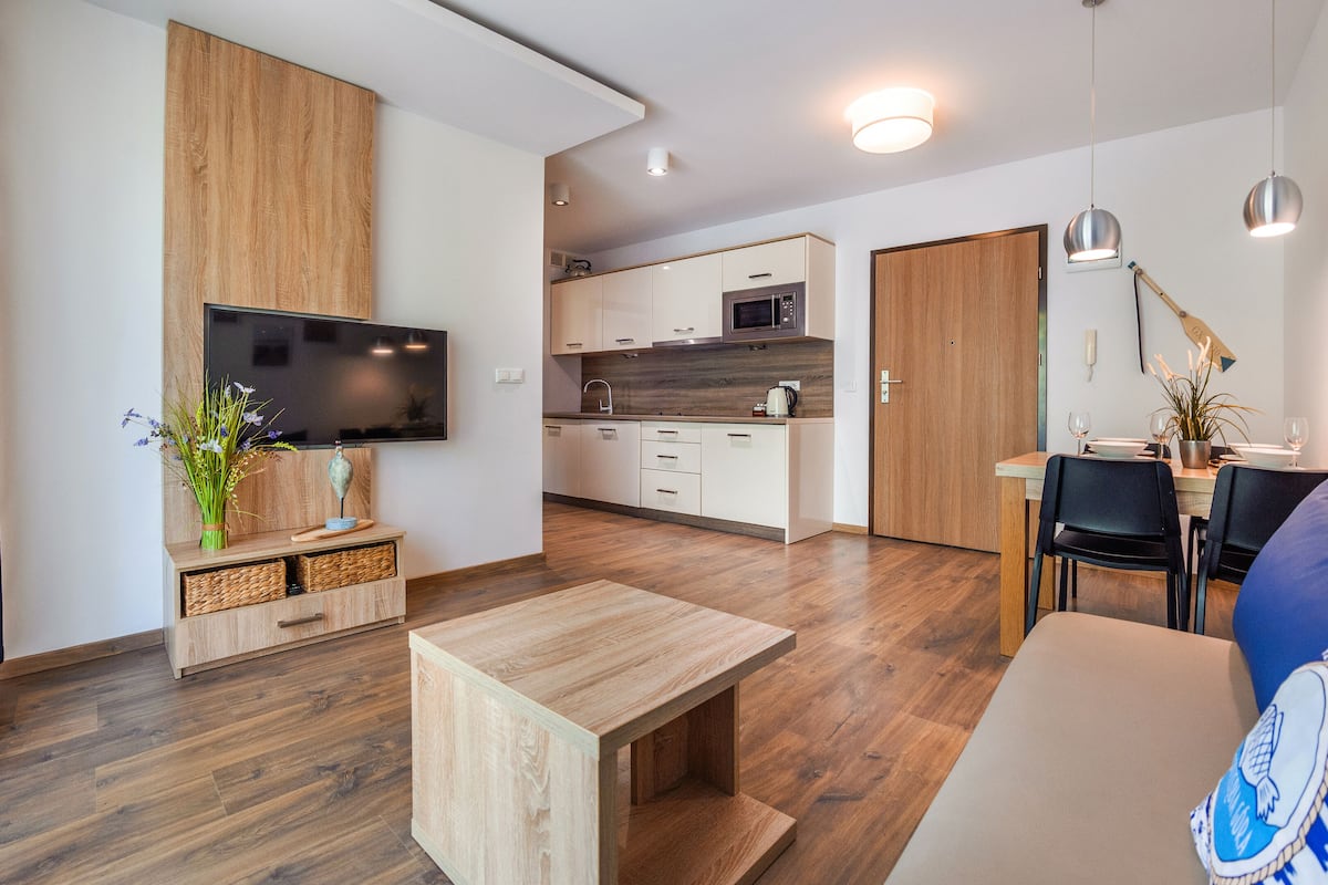 Ein modernes Wohnzimmer mit Couchtisch aus Holz, einem blau gestreiften Sofa im Marine-Stil, einem an einer Holzwand montierten Flachbildfernseher und einer Küchenzeile mit Mikrowelle und Schränken.