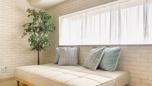 A modern room with a white brick wall, a large window with sheer curtains, a beige sofa with patterned cushions, and a green plant. - Shioji Aoi Villa (Osaka)