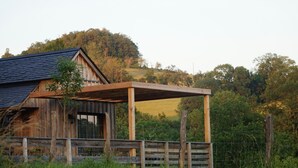 A rustic wooden house with a covered porch, surrounded by lush greenery and hills in the background.