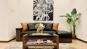 A modern living room with a black leather sofa, a glass coffee table with magazines, a decorative vase with flowers, and a large black and white abstract painting on the wall. - Asahi Kasumi Villa (Osaka)