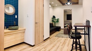 A compact living space with a wooden floor, a blue-tiled wall, a wooden door, a ceiling fan, a small dining area with stools, and a sofa. - Asahi Kasumi Villa (Osaka)