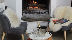 Un salón moderno con chimenea, dos sillones grises con mantas de piel blanca, un libro, una botella de champán y copas sobre una bandeja.