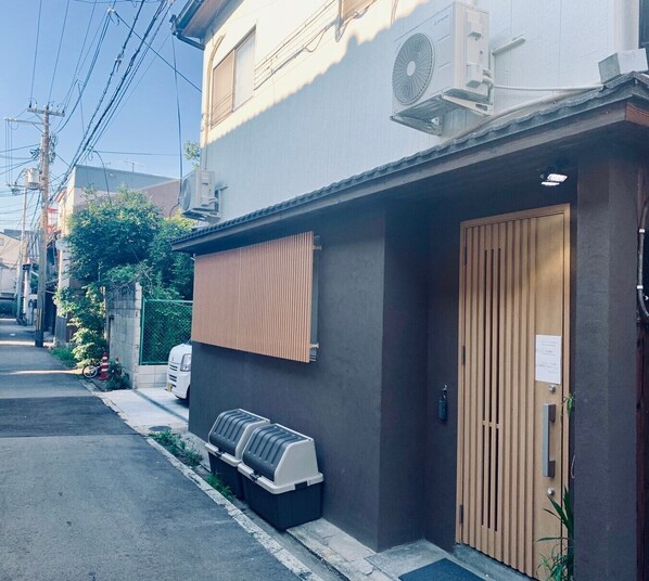 A residential building with a wooden door, a small outdoor grill, and a wall-mounted air conditioning unit. - Hanazonominami Apartment 101 (Osaka)