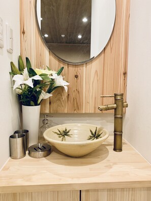 A bathroom with a wooden vanity, a round mirror, a vessel sink, and a faucet. There is a vase with flowers on the vanity. - Hanazonominami Apartment 101 (Osaka)