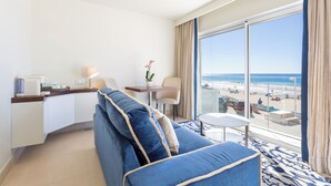 A hotel room with a large window offering a view of the beach, a blue sofa, a small round table, and a kitchenette area.