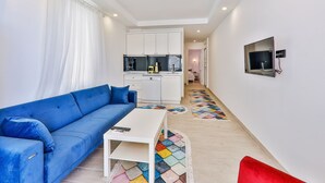 Un espacio habitable compacto con un sofá azul, una mesita blanca, una zona de cocina y un televisor fijado a la pared.