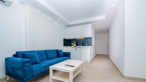Un espacio habitable pequeño con un sofá azul, una mesita de centro blanca y una kitchenette con armarios blancos y una encimera negra.