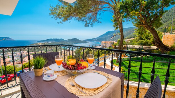 Mesa de balcón con frutero, vasos y un plato blanco, con vista a un paisaje costero.