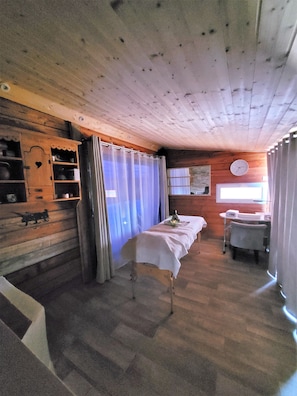 Interior de una cabaña de madera con una cama, una mesita y un estante con objetos.