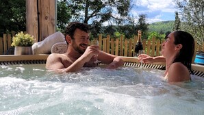 Un couple dans un jacuzzi, une bouteille de champagne à portée de main, entouré de clôtures en bois et de verdure.