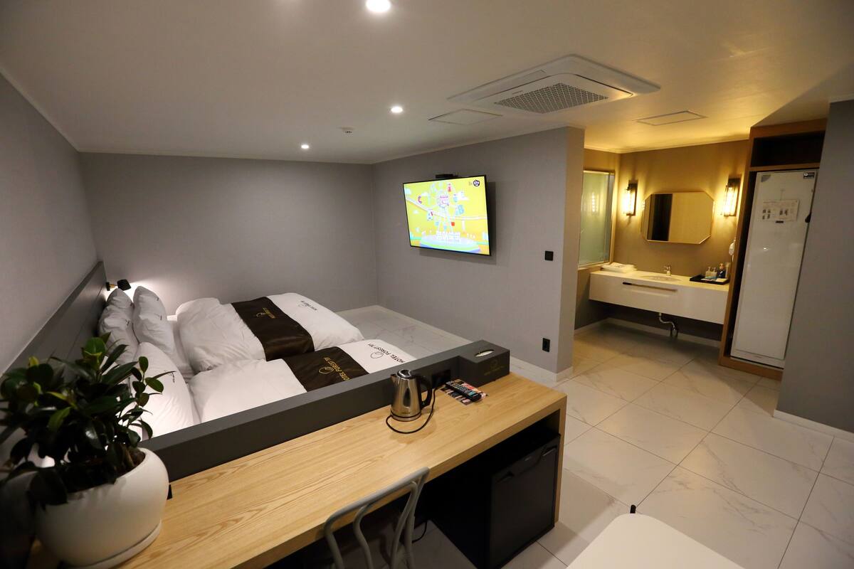 A hotel room with a bed, a desk with a plant, a TV, a bathroom with a mirror, and a refrigerator.