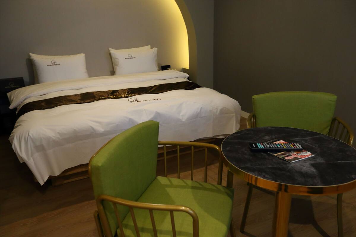 A neatly made bed with white linens and two pillows, a green upholstered chair, a small round table with a remote control and magazine, and a wooden floor.
