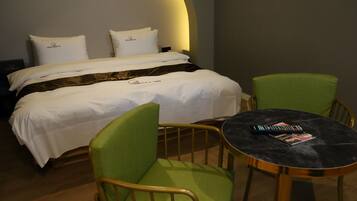 A neatly made bed with white linens and two pillows, a green upholstered chair, a small round table with a remote control and magazine, and a wooden floor.