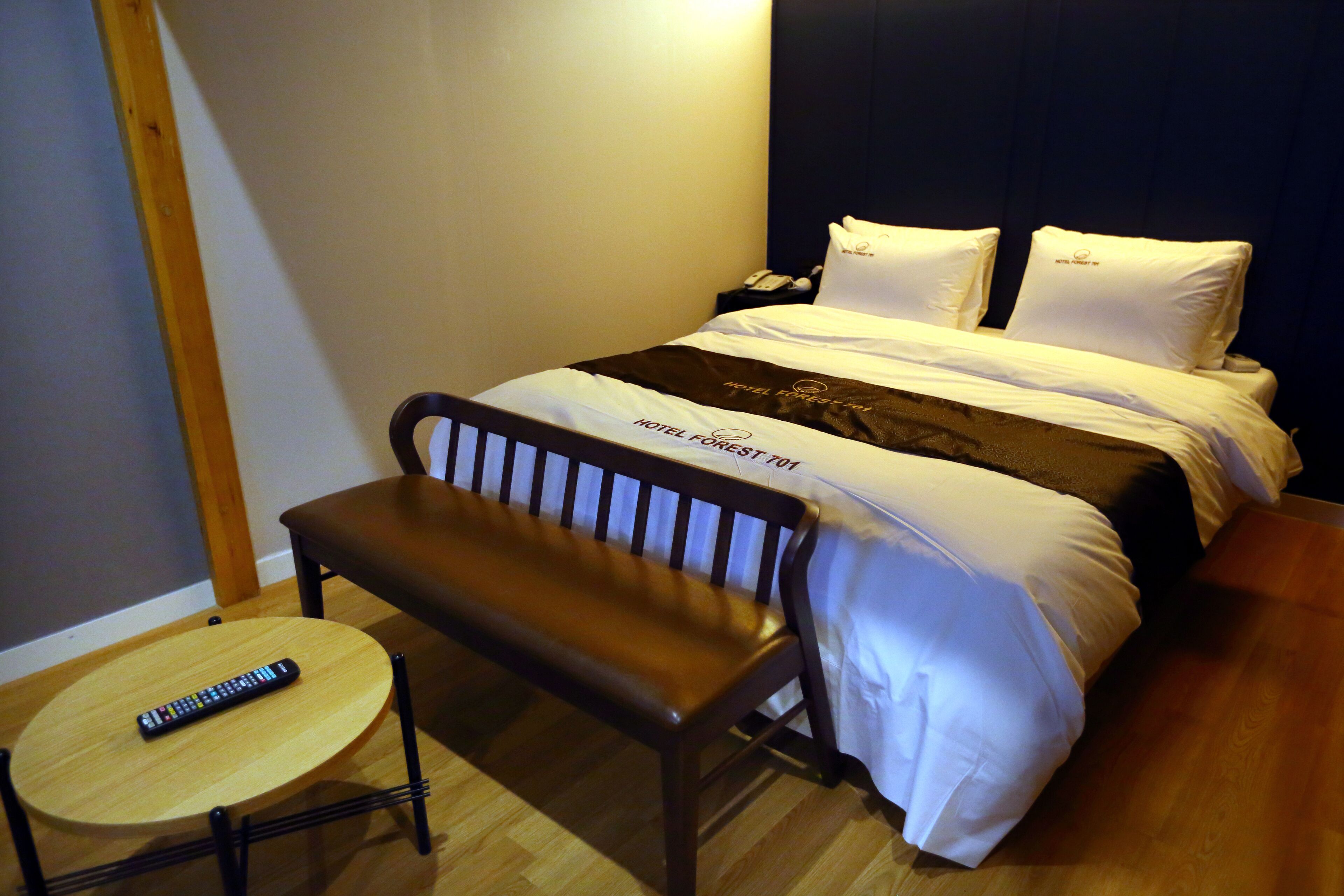 A hotel room with a bed, a bedside table, a remote control, and a bench.