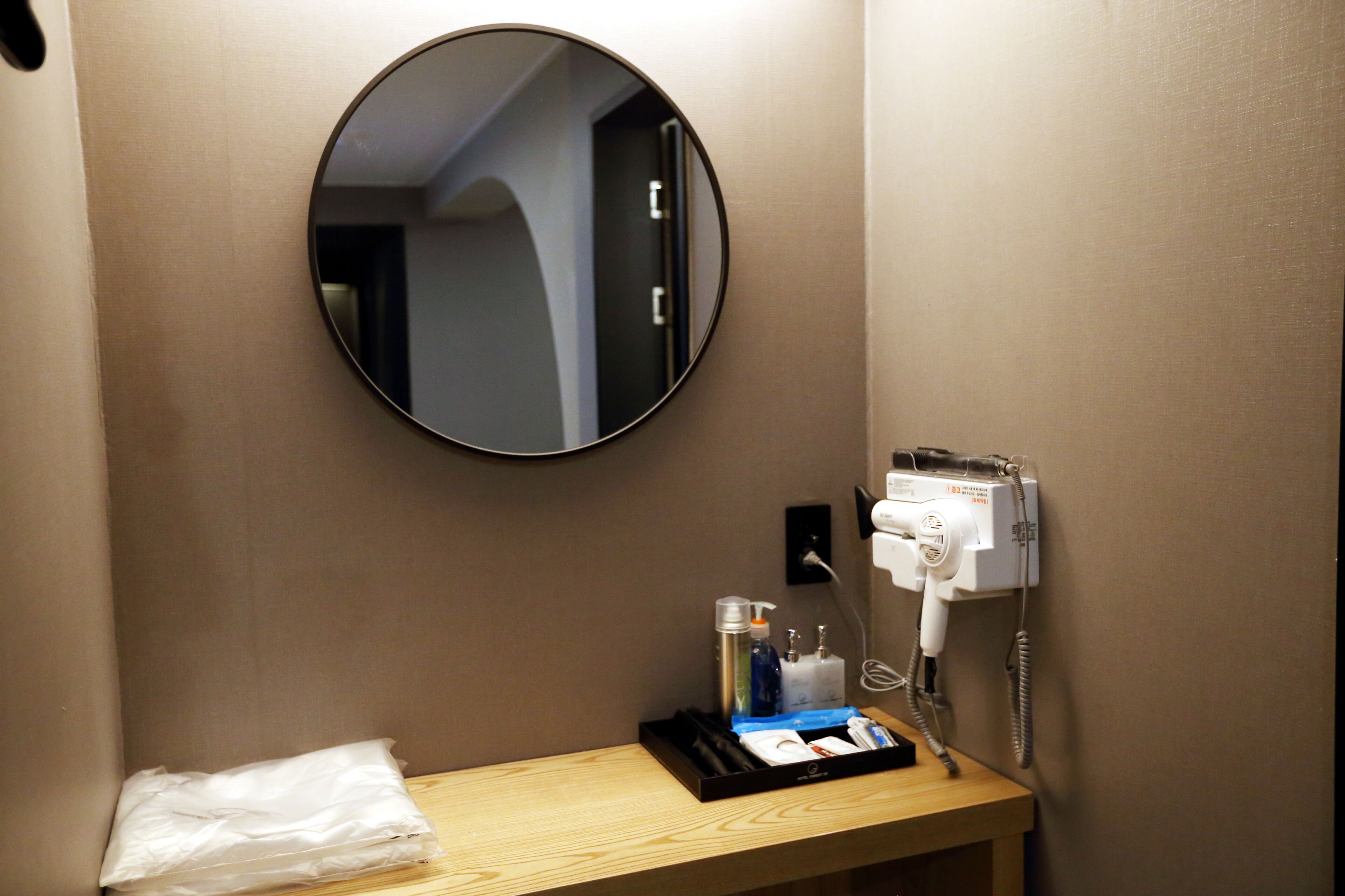 Une salle de bain d’hôtel avec un miroir rond, un sèche-cheveux et des articles de toilette disposés sur un comptoir en bois.