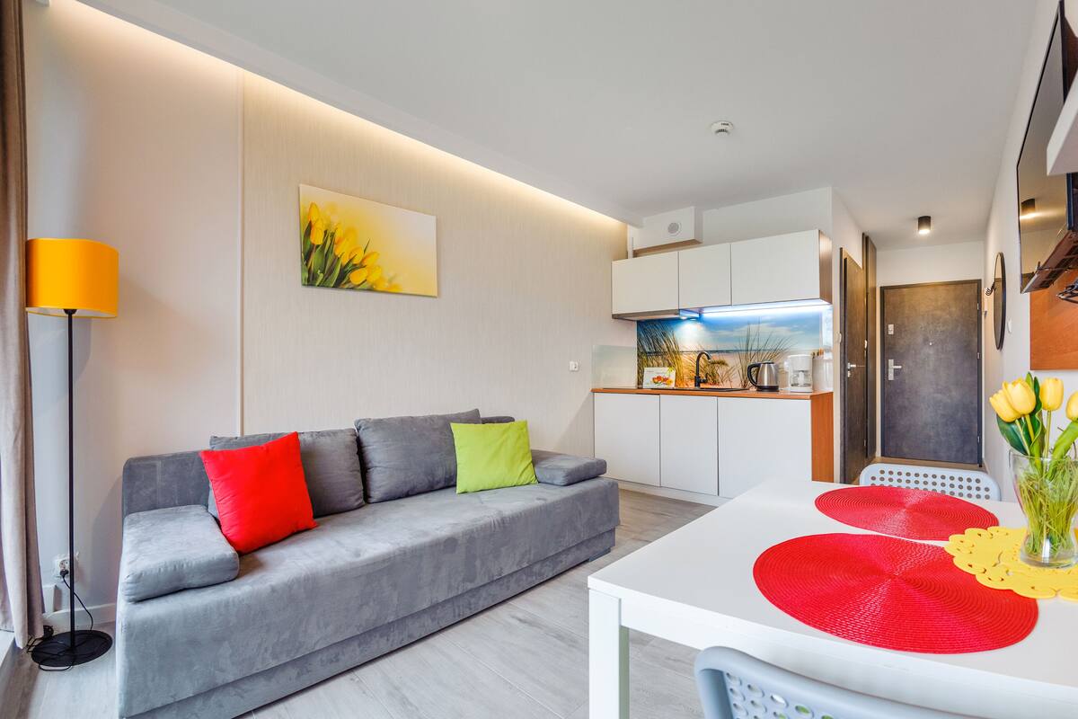 A modern living room with a grey sofa, a dining table with red placemats, and a kitchen in the background.