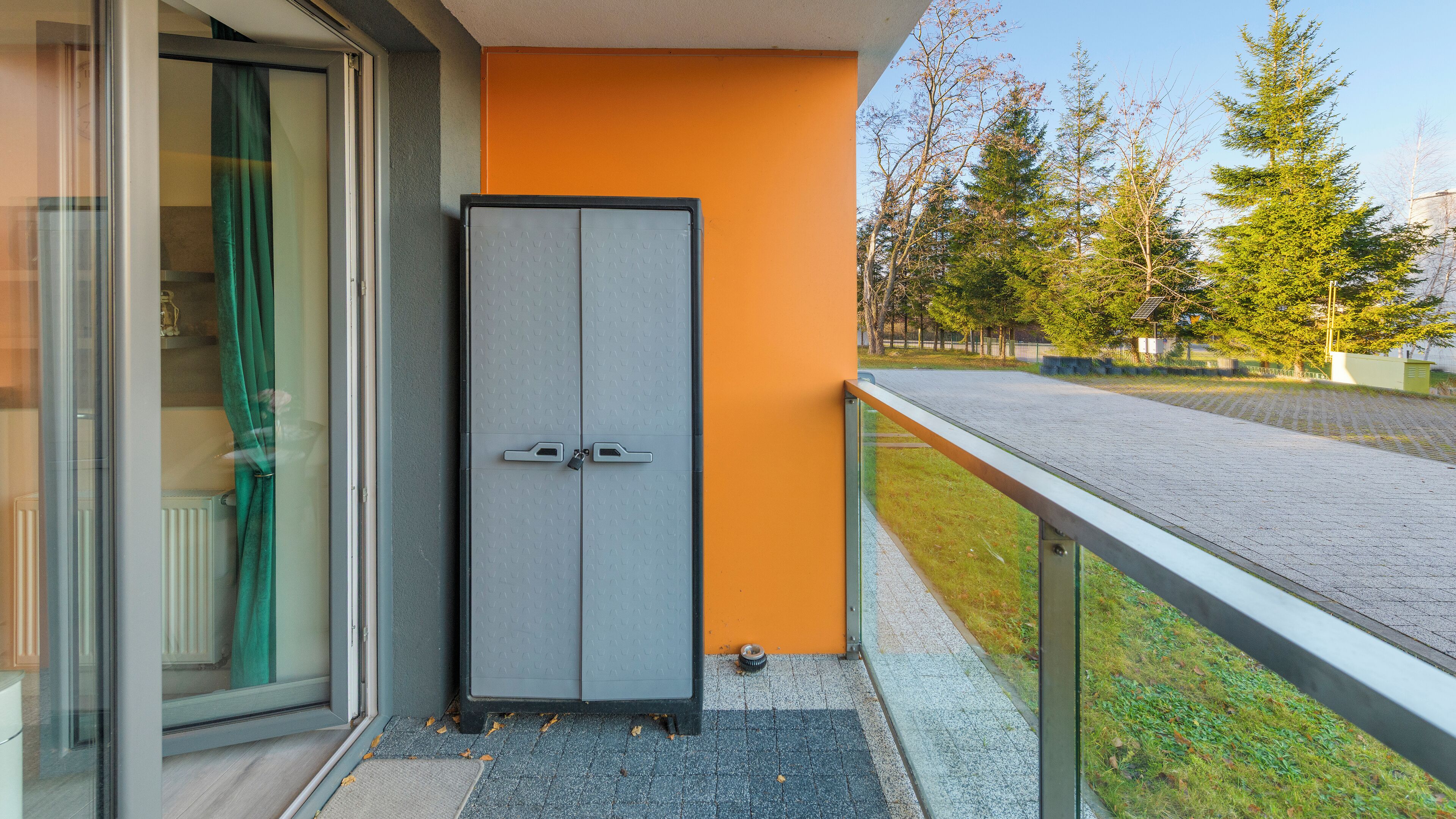 A modern building with a grey door and an orange wall, a metal railing, and a paved area with trees in the background.