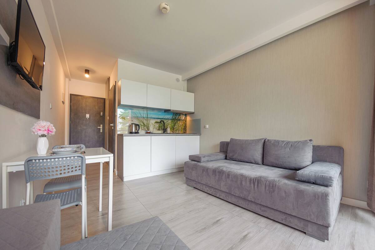 A modern living room with a grey sofa, a dining table with chairs, and a kitchen area visible through an open door.