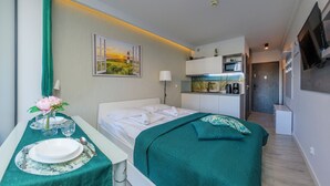 A hotel room with a bed, a desk with a vase of flowers, a painting of a sunset, a kitchenette, and a TV mounted on the wall. - Apartamenty Sun & Snow Sanatoryjna (Wladyslawowo)