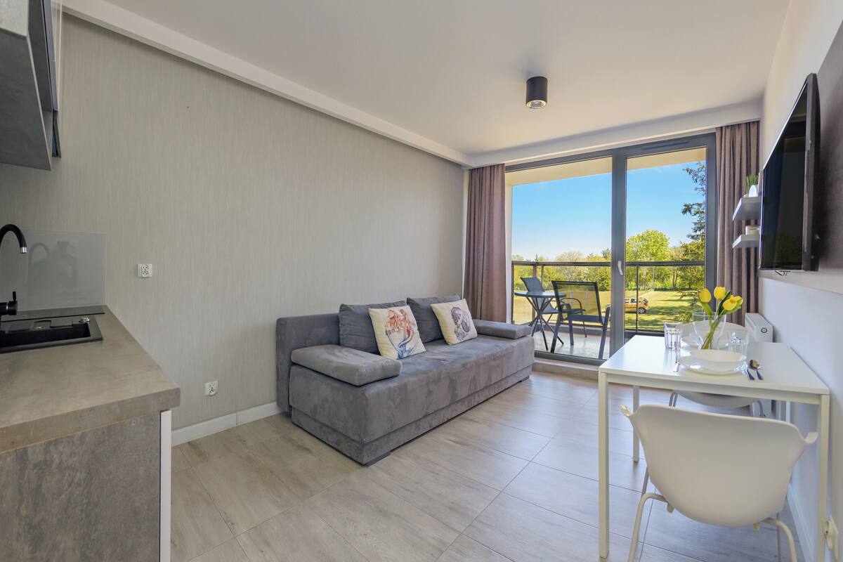 A modern living room with a grey sofa, a dining table with chairs, a TV mounted on the wall, and a balcony with a view of the outdoors.