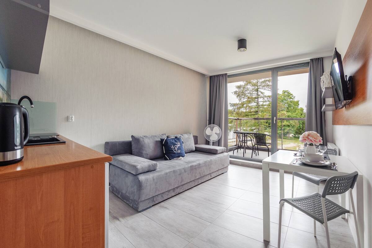 A modern living room with a grey sofa, a wooden desk, a small table with a chair, and a balcony with a view of trees.