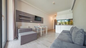A modern living room with a grey sofa, a dining table with chairs, a TV mounted on the wall, and a kitchen area with cabinets and a window. - Apartamenty Sun & Snow Sanatoryjna (Wladyslawowo)