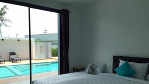 A hotel room with a large sliding glass door leading to a pool area, a bed with a striped bedspread, and a nightstand with a lamp. - The Phu Resort (Pranburi)