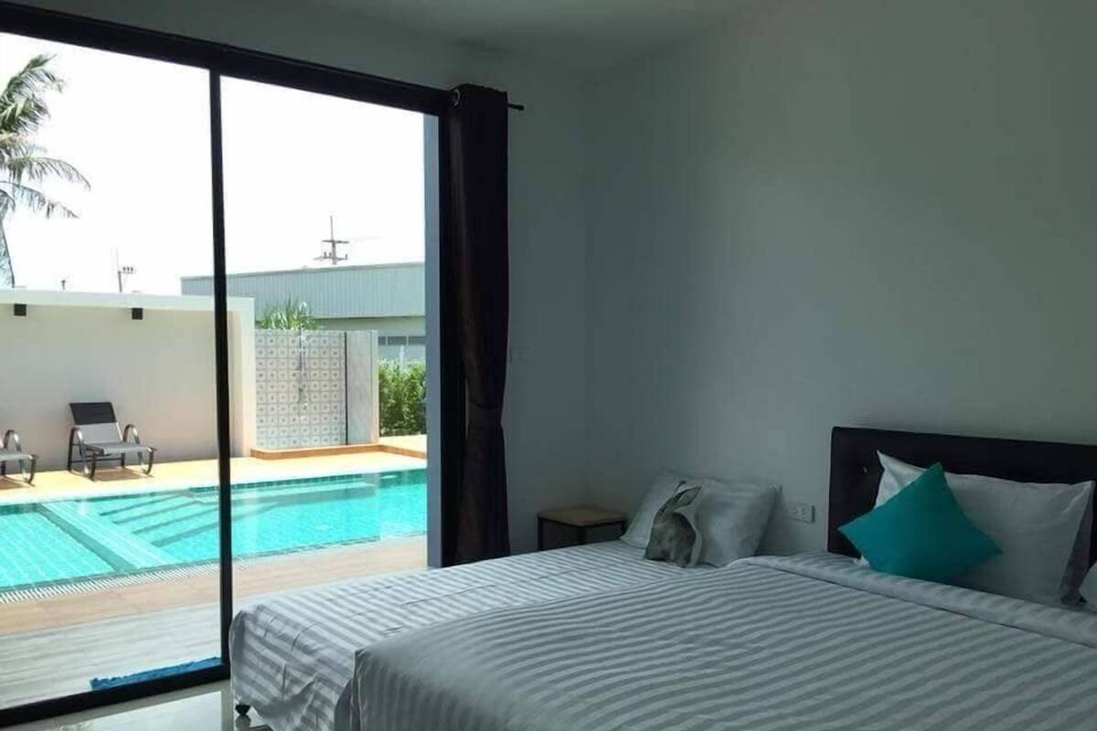 Quarto de hotel com uma grande porta de vidro deslizante que dá para uma área de piscina, uma cama com colcha listrada e uma mesinha de cabeceira com abajur.