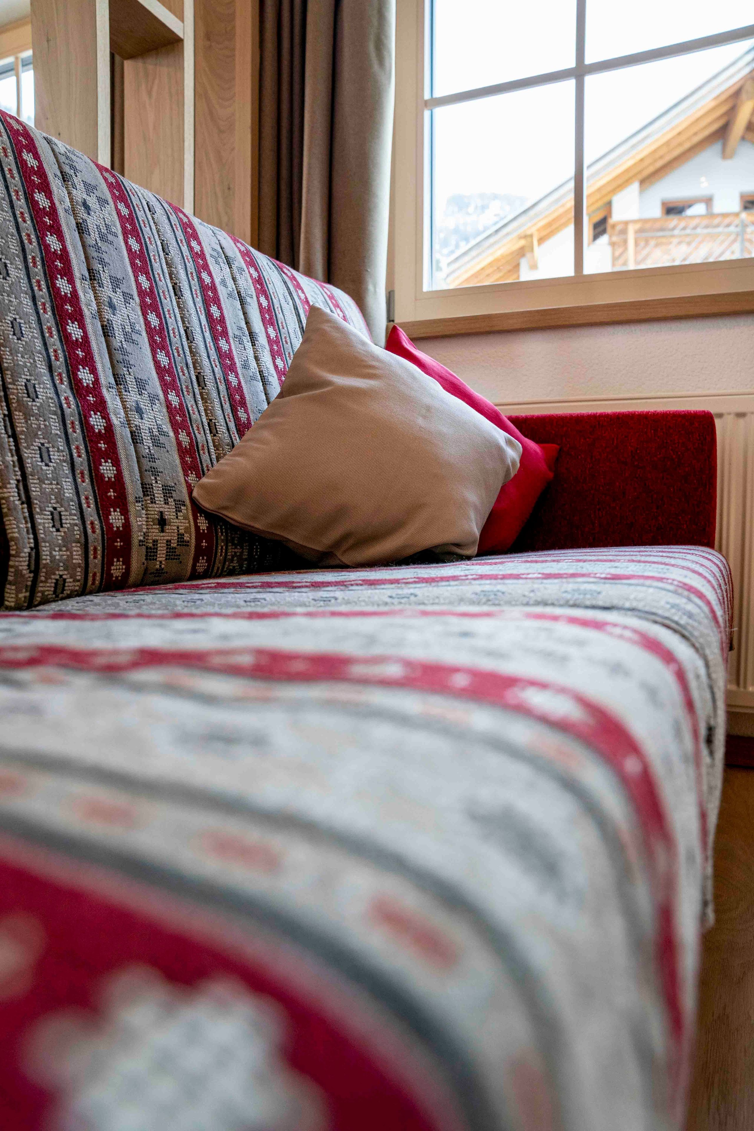 A bed with patterned bedding, a red pillow, and a window with a view of a wooden structure.