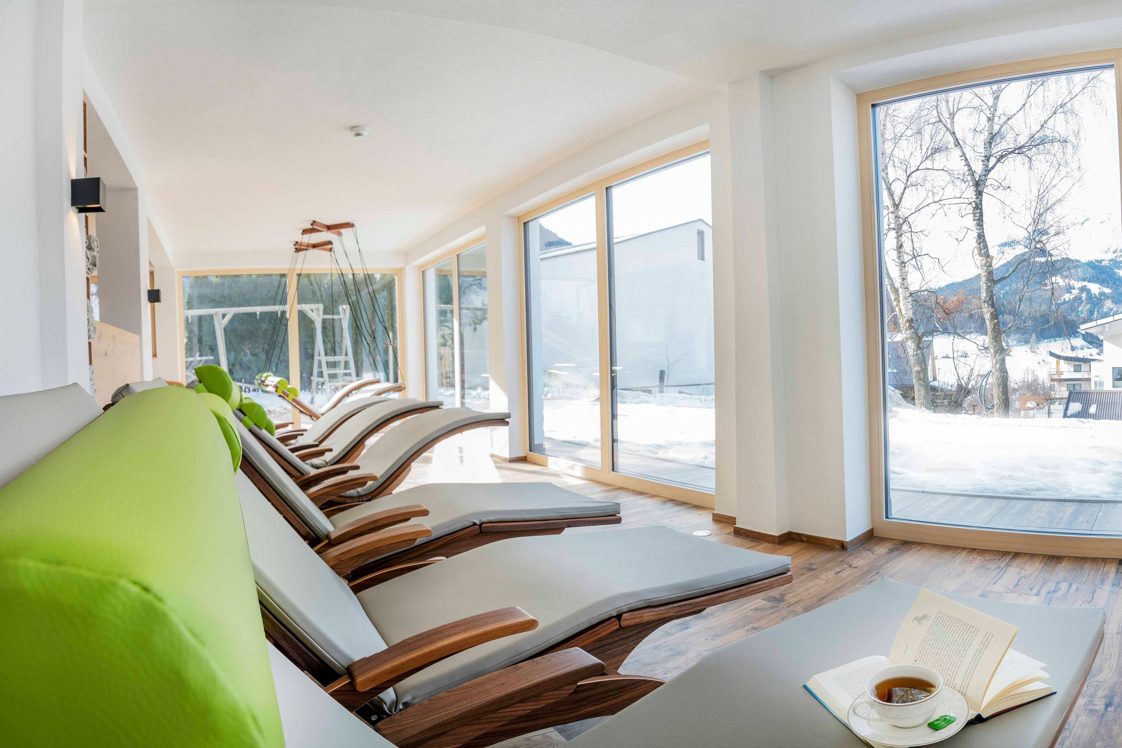 A modern spa room with wooden loungers, a reading area with a book and cup, and large windows with a snowy view.