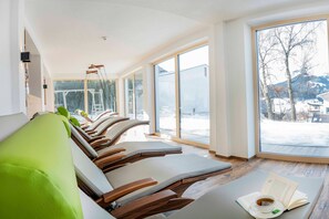 A modern spa room with wooden loungers, a reading area with a book and cup, and large windows with a snowy view.