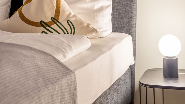 Una cama bien tendida con cabecera gris, almohadas blancas y doradas, y una mesita de noche con lámpara.