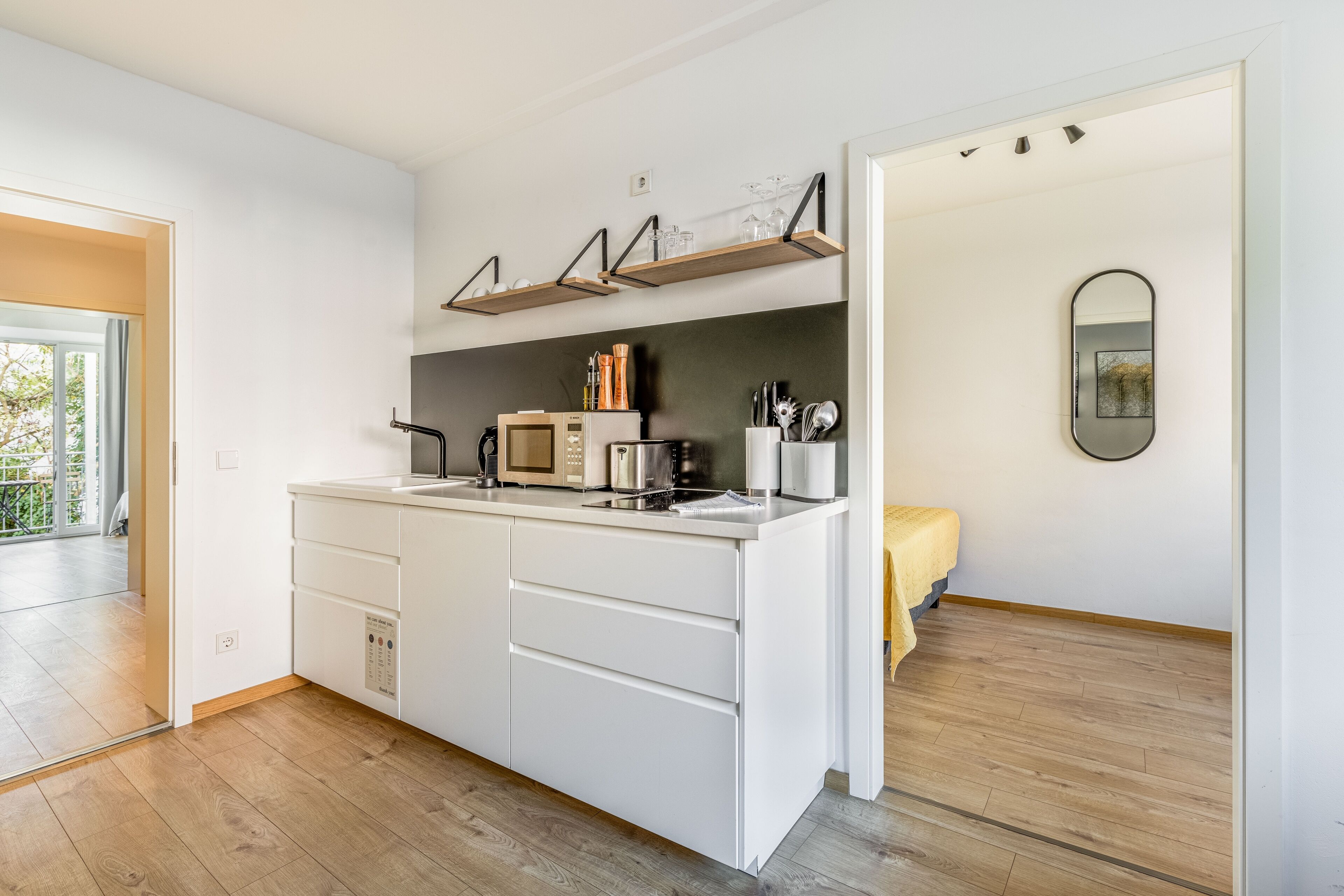 A modern kitchen with white cabinetry, a built-in microwave, and a coffee machine. There are wooden shelves above the counter and a small mirror on the wall.