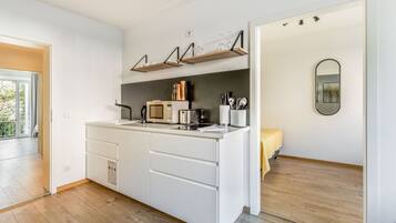 A modern kitchen with white cabinetry, a built-in microwave, and a coffee machine. There are wooden shelves above the counter and a small mirror on the wall.