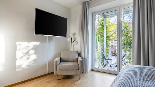 A modern hotel room with a large flat-screen TV mounted on the wall, a grey armchair, a bed with a grey patterned cover, and a balcony with a view of greenery and buildings. - Limehome Augsburg Pilgerhausstraße (Augsburg)