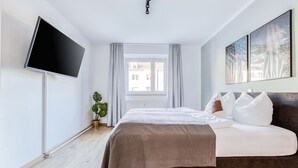 A modern bedroom with a large bed, a flat-screen TV mounted on the wall, and a window with curtains. - Limehome Augsburg Pilgerhausstraße (Augsburg)