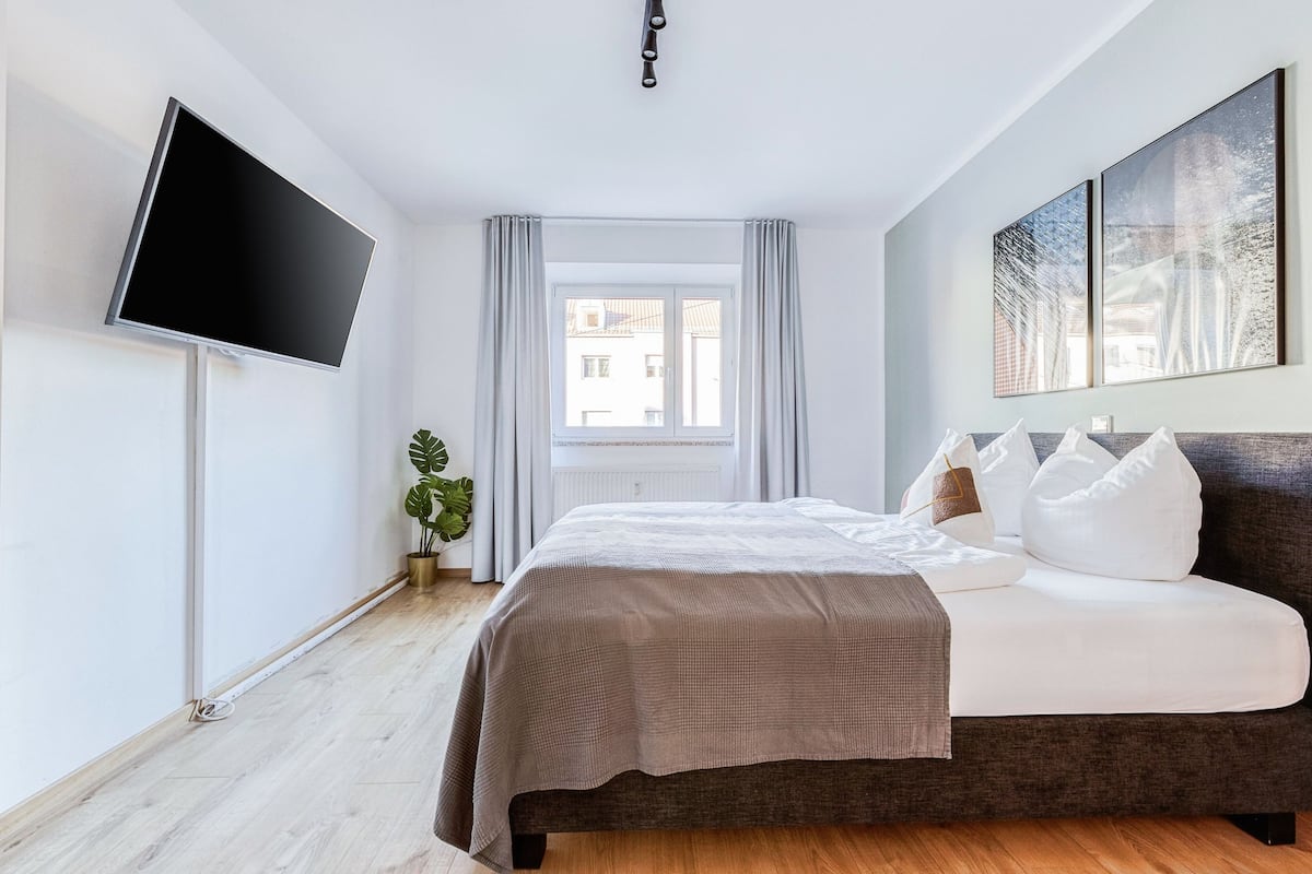 A modern bedroom with a large bed, a flat-screen TV mounted on the wall, and a window with curtains.