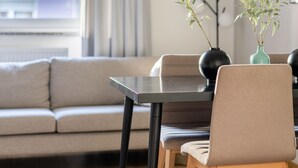 A modern living room with a sofa, a dining table with chairs, a vase with plants, and a window with curtains. - Limehome Augsburg Pilgerhausstraße (Augsburg)