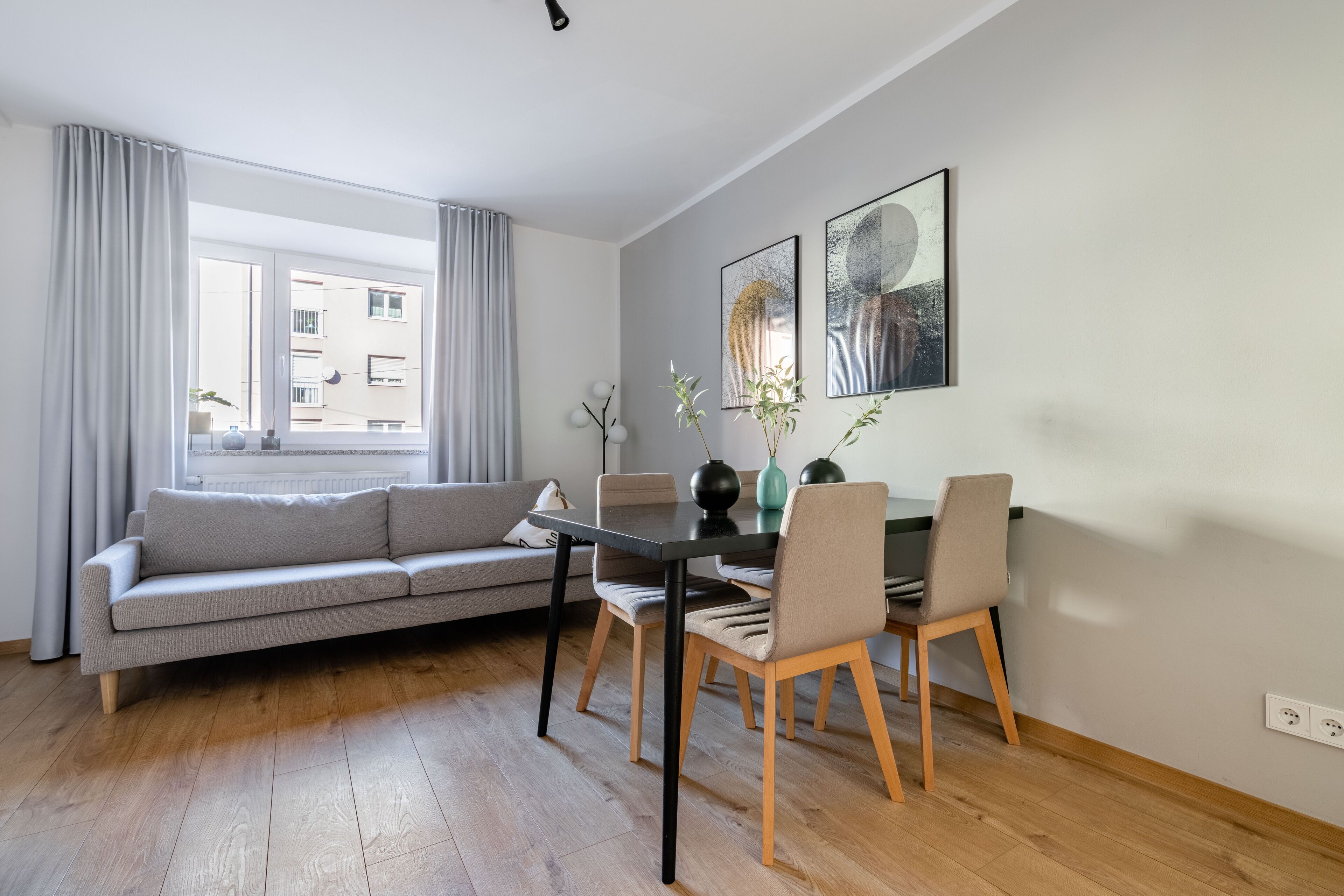 Una sala moderna con un sofá gris, una mesa de comedor de madera y sillas. Hay dos cuadros enmarcados en la pared y una ventana con cortinas.