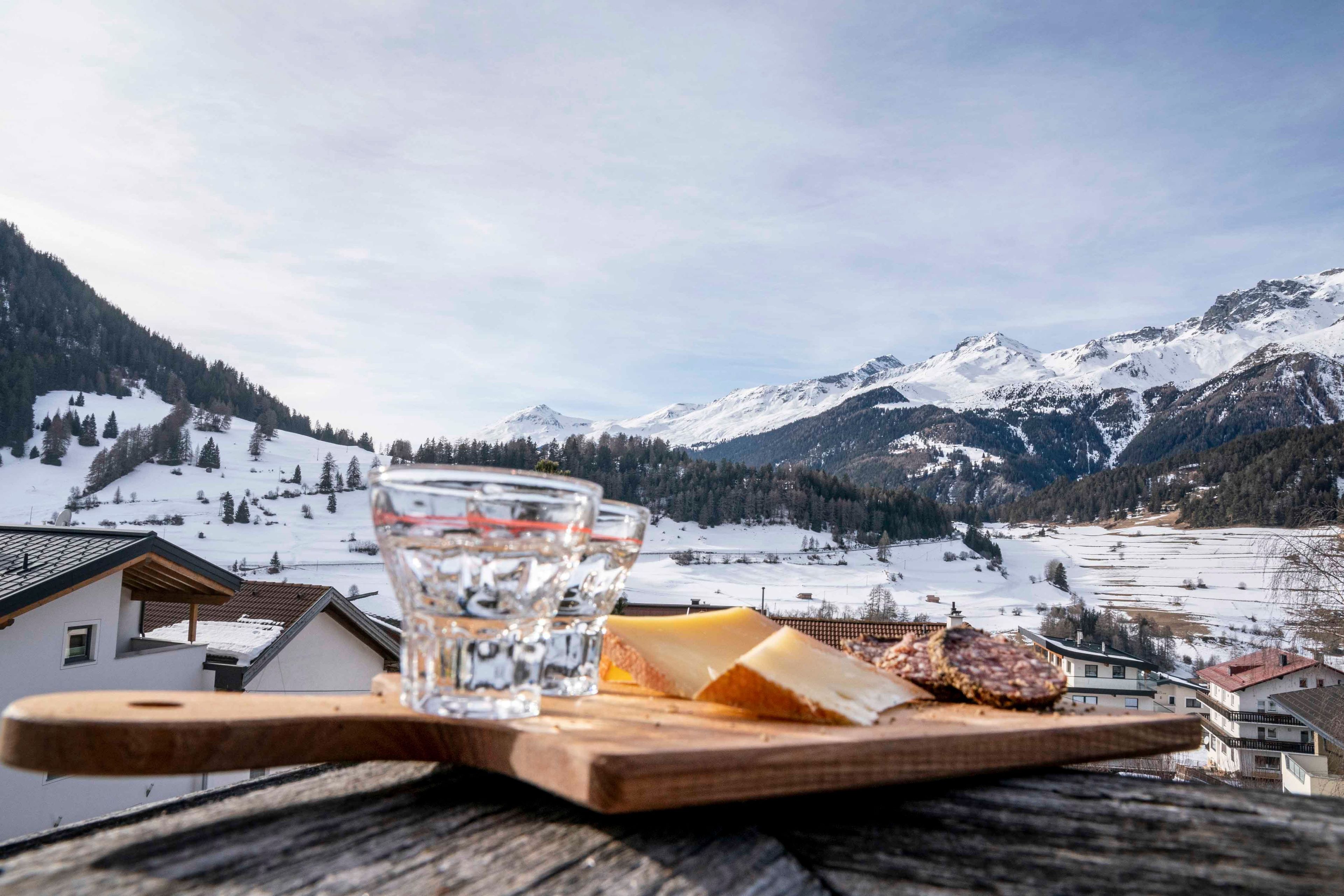 Une planche à découper en bois, un verre d’eau, du fromage et une tranche de viande sont posés sur une table, avec une vue sur les montagnes en arrière-plan.